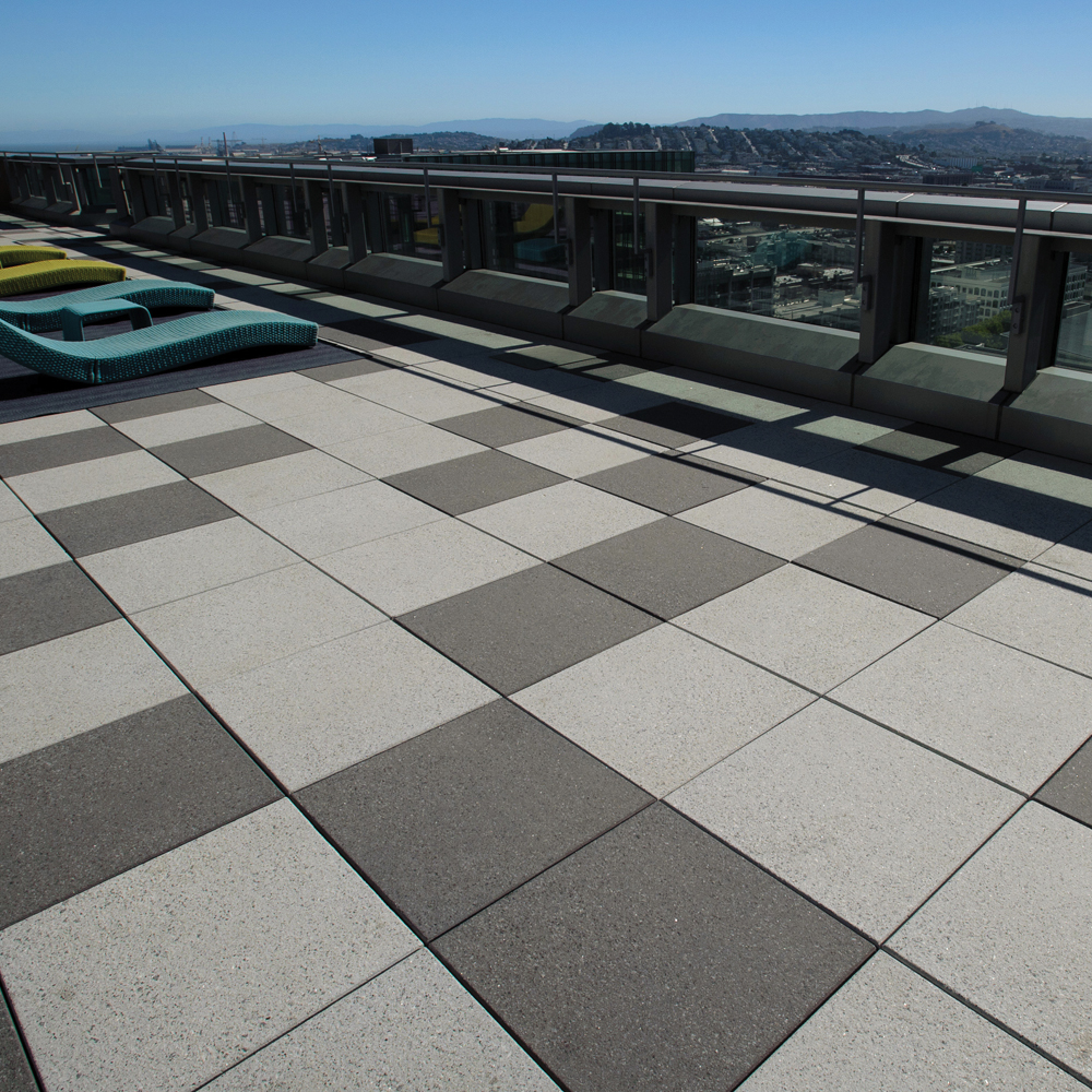 This outdoor area has white and gray square pavers. There are blue and green lounge chairs looking out to the city and mountain view.