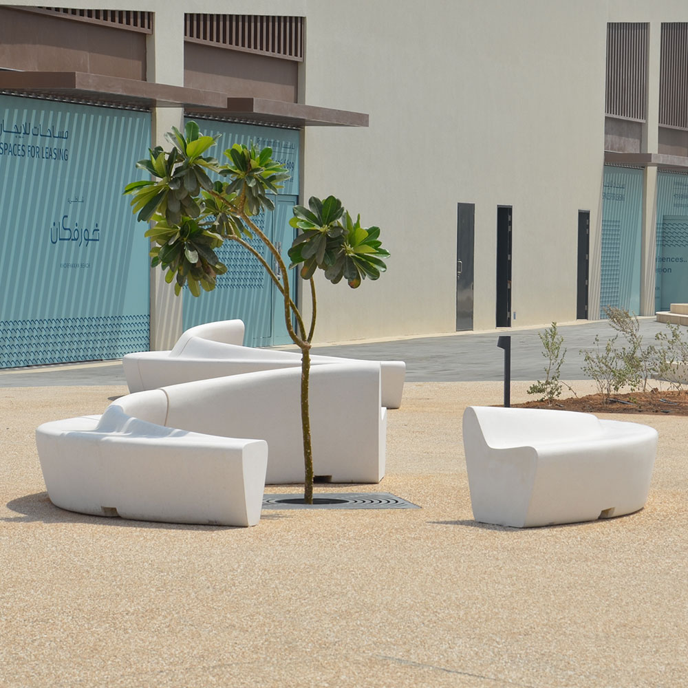 An outdoor seating area with modern-style, asymmetrical white concrete benches. A small tree sits in the middle.