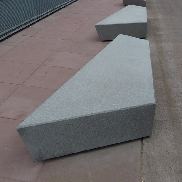 A light-gray, asymmetrical concrete bench in a walkway with red pavers.