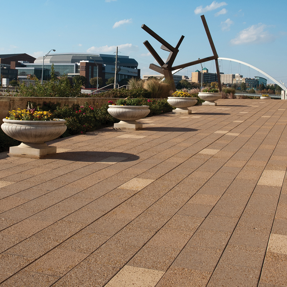 A wide outdoor walkway with a dark brown abstract sculpture, a city skyline and a large bridge in the background. Concrete flower pots line the light brown tiles.