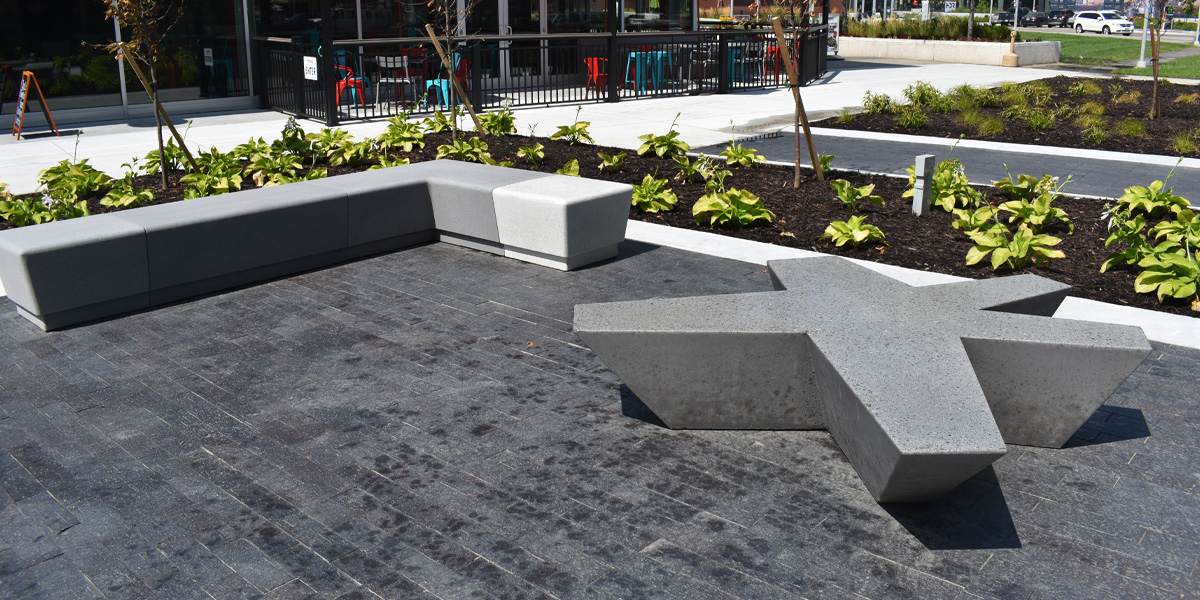 An asterisk-shaped concrete bench in a plaza with dark gray pavers and small bushes in the background.