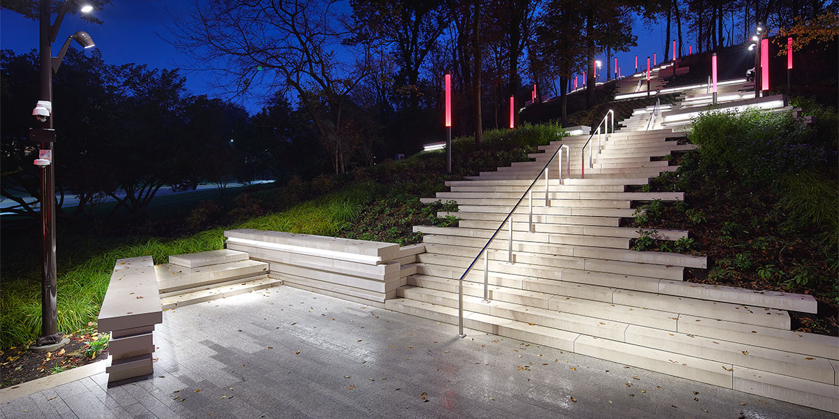 An asymmetrical, white, outdoor stairway with vertical red lights lining it. An illuminated silver railing sits in the middle.