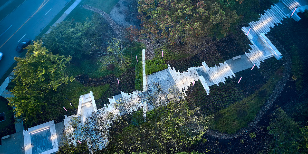 An aerial view of an asymmetrical, white, outdoor stairway resembling a soundwave. Green treetops surround it and a two-way road in the top left corner.