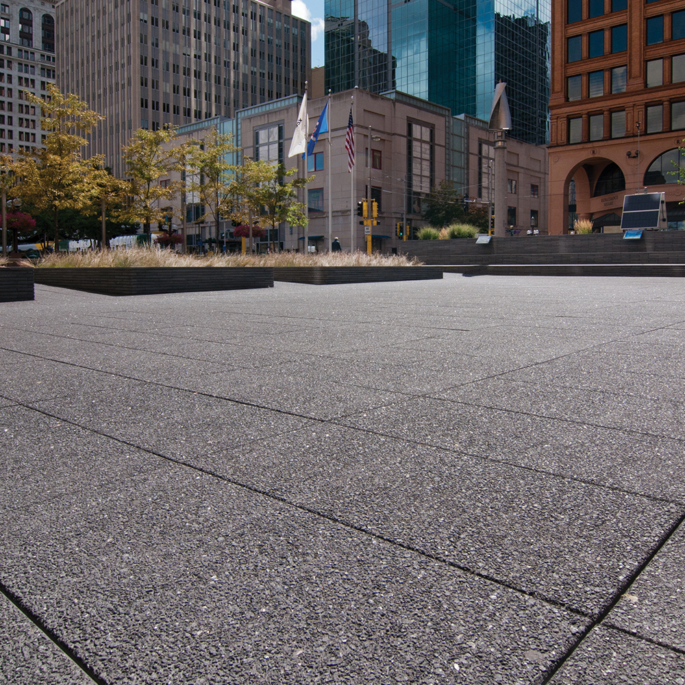 This city has a large area with black, speckled pavers. There are three flags in the distance with various buildings.