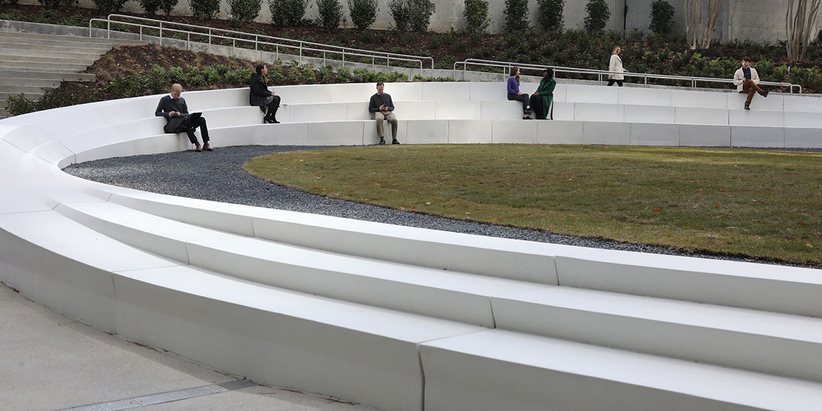 A round grass plaza surrounded by a concrete barrier with three steps. People are sitting on the barrier facing the grass.