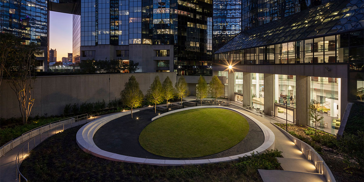 A round grass plaza at dusk with a concrete sidewalk surrounded by skyscrapers on every side. Six trees line the inside edge.