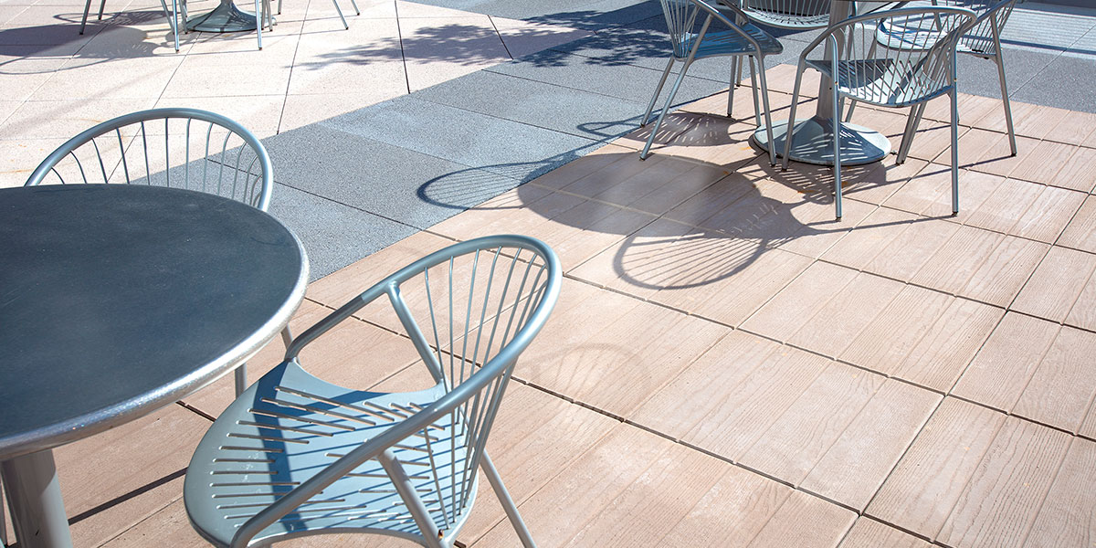 A plaza balcony with round stainless steel tables surrounded by stainless steel chairs. The ground Is paved with gray and tan tile.