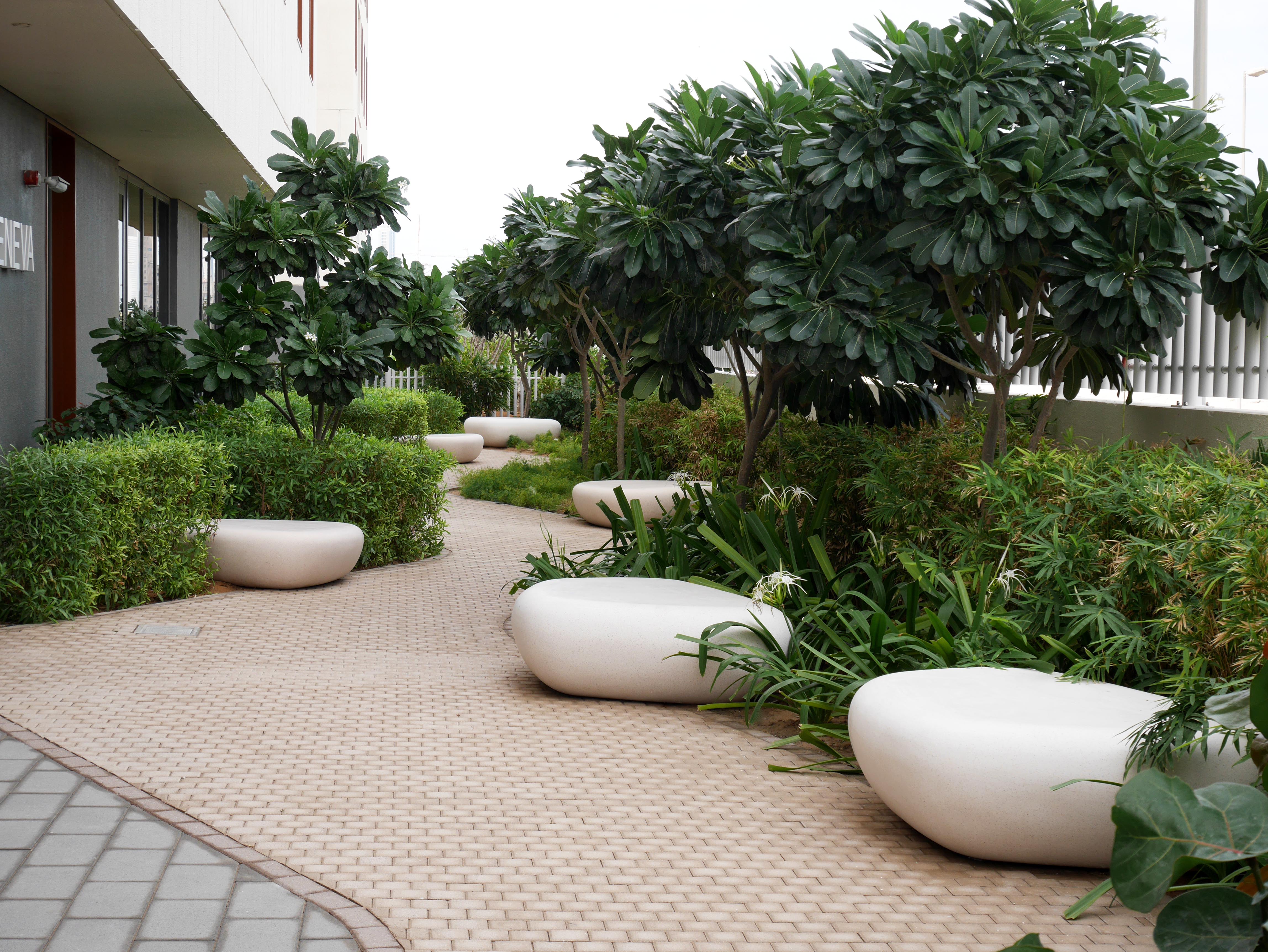 A beige brick walkway surrounded by trees and bushes. Bean shaped concrete benches line the walkway.