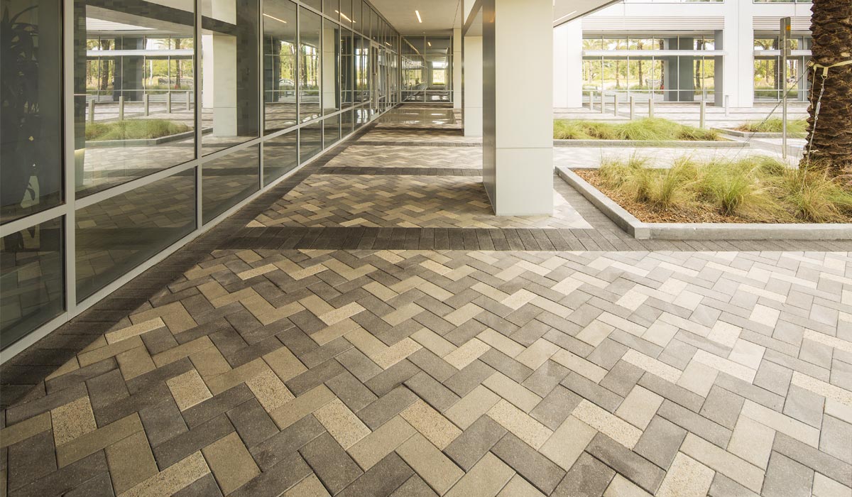 A courtyard of a business park with beige and gray pavers in a zig-zag pattern. The perimeter of the courtyard is covered to make a walkwayn.