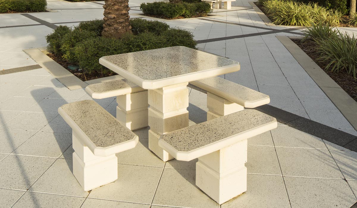 A concrete seating area with four benches surrounding a square table in a courtyard with white and dark gray pavers.