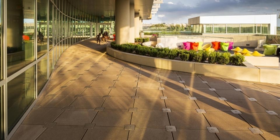 A roof deck with square, brown concrete tile secured with Wausau Tile's Lok-Down system. A seating area with orange, pink, and green furniture is enclosed with a concrete median landscaped with bushes.